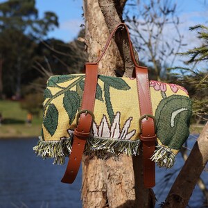 May include: A picnic blanket with a floral design in yellow, green, and pink, featuring brown leather straps and buckles. The blanket has a fringed edge and is hanging on a tree branch.