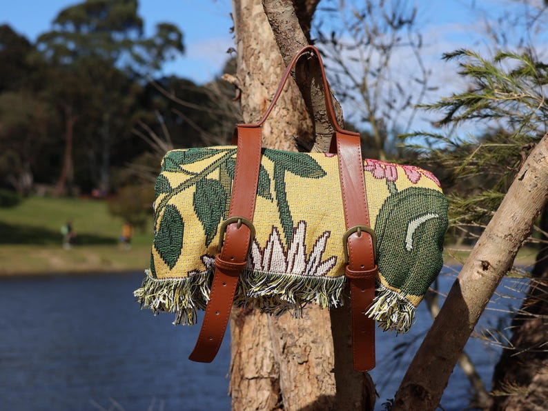 May include: A picnic blanket with a floral design in yellow, green, and pink, featuring brown leather straps and buckles. The blanket has a fringed edge and is hanging on a tree branch.