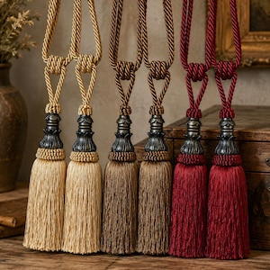 Pode incluir: Seis braçadeiras decorativas para cortinas dispostas em pares de dourado, bronze e vermelho profundo. Cada braçadeira apresenta um laço de corda torcida, uma tampa ornamental metálica escura com detalhes intrincados e uma borla densa.