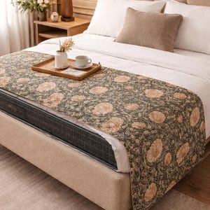 May include: A bed with a beige upholstered frame and a patterned bed runner. The runner features a floral design in shades of green, beige, and peach. A wooden tray with a cup and vase sits on the bed. A beige pillow and white sheets complete the bedding.