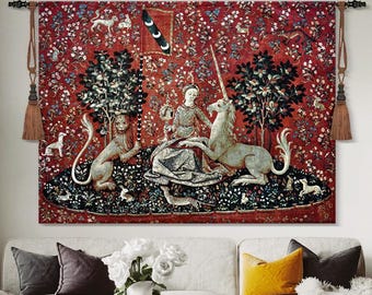 May include: A large tapestry featuring a medieval scene with a woman, a unicorn, and a lion. The artwork has a deep red background with floral and animal patterns. The tapestry is suspended on a wall with decorative tassels.