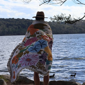 May include: A person wearing a brown hat and a floral patterned blanket stands near a lake. The blanket features large flowers in shades of orange, pink, blue, and purple. The blanket has a white fringe.