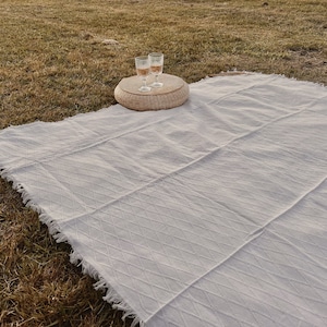May include: A white picnic blanket with a fringed edge is spread on a grassy surface. Two glasses of a light-colored beverage sit on a small, round woven table. The blanket has a textured, diamond pattern.