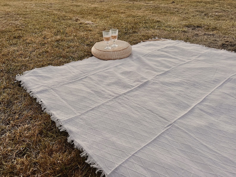May include: A white picnic blanket with a fringed edge is spread on a grassy surface. Two glasses of a light-colored beverage sit on a small, round woven table. The blanket has a textured, diamond pattern.