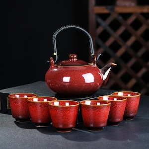 Może przedstawiać: Czerwony ceramiczny zestaw do herbaty z pasującym imbrykiem i sześcioma małymi filiżankami. Czajnik ma czarny uchwyt i dzióbek. Filiżanki i czajnik mają nakrapiany wzór ze złotym wykończeniem. Zestaw jest do herbaty.