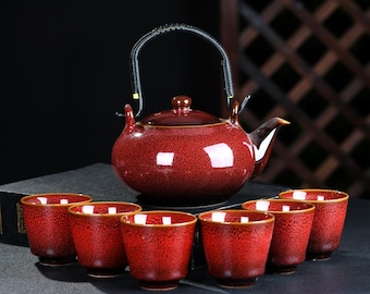 May include: A red ceramic tea set with a matching teapot and six small cups. The teapot has a black handle and a spout. The cups and teapot have a speckled design with gold trim. The set is designed for tea.