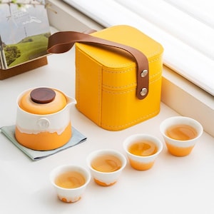 Puede incluir: Un juego de té amarillo con un estuche de transporte a juego. El juego incluye una tetera con tapa de madera, cuatro tazas de té pequeñas y un estuche cuadrado con asa marrón. Las tazas y la tetera tienen detalles en naranja y blanco.