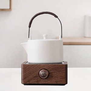 Puede incluir: Tetera de cerámica blanca con asa de cuero marrón, sentada sobre una base de madera con una perilla de cobre. La base tiene un cable para uso eléctrico.