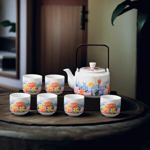 May include: A white ceramic tea set with a teapot and six cups, adorned with vibrant floral patterns in orange, yellow, blue, and pink. The teapot has a black handle and a lid with a similar floral design. The set is designed for serving tea.