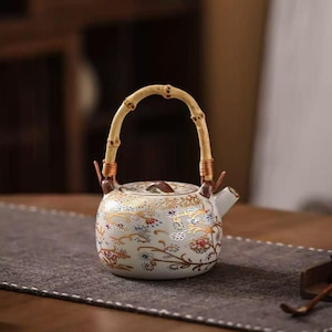 Puede incluir: Una tetera de cerámica blanca con un asa de bambú y un estampado floral en dorado, rojo y azul. La tetera tiene un pequeño pico y una tapa con un pomo marrón. El asa está sujeta con alambre de cobre.
