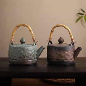 Op de afbeelding: Twee keramische theepotten met bamboe handvatten. Eén is gevlekt groen, de andere bruin. Beide hebben een tuit en een deksel met een kleine knop. De handvatten zijn bevestigd met koperdraad.