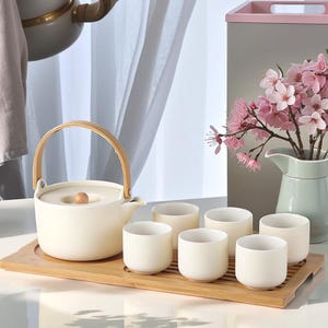 May include: A white ceramic teapot with a wooden handle and lid, along with six white ceramic teacups, all arranged on a bamboo tray.