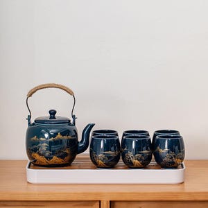 May include: A dark teal ceramic tea set comprising a teapot and four cups. The teapot has a rope handle and a lid. The set features a gold landscape design. The set is displayed on a white tray.