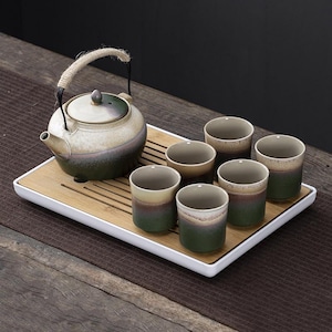 Puede incluir: Un juego de té de cerámica con una tetera y seis tazas a juego. La tetera tiene un asa de cuerda y una tapa. Las tazas y la tetera tienen un esquema de color degradado de verde, beige y marrón. El juego está sobre una bandeja de bambú con borde blanco.