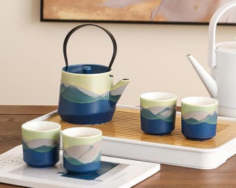 May include: A ceramic tea set with a blue, green, and yellow mountain design. The set includes a teapot with a black handle and four matching teacups. The tea set is displayed on a bamboo tray and a white tray.