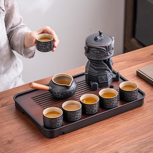 May include: A black ceramic tea set with a distinctive design, including a teapot with a wooden handle, a tea dispenser, and six small cups. The set is arranged on a dark wooden tray. One cup is being held.