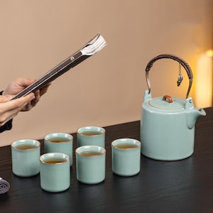 May include: A light blue ceramic tea set with six matching cups, a teapot, and a book. The teapot has a brown handle and a small charm. The cups and teapot are filled with tea.