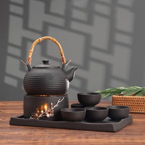 Könnte beinhalten: Schwarzes Keramik-Teeservice mit Bambusgriff an der Teekanne. Das Set umfasst eine Teekanne, einen Wärmer und mehrere kleine Tassen, die alle auf einem rechteckigen Tablett angeordnet sind. Der Wärmer hat ein Blumendesign.