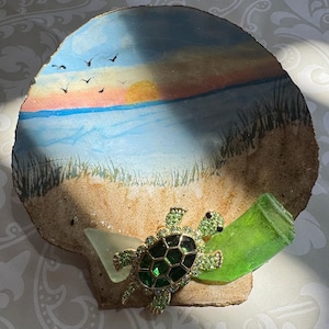 May include: A circular decorative shell painted with a beach sunset scene featuring a yellow sun, blue water, and sand dunes. A green sea turtle figurine with a dark green shell and light green accents is attached, flanked by two pieces of sea glass.