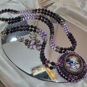 Op de afbeelding: Een meerstrengs ketting met een grote, ronde hanger met een vrouwen gezicht met een blauw oogmasker. De ketting is gemaakt van paarse en groene kralen en zilveren accenten. Passende oorbellen zijn ook inbegrepen.