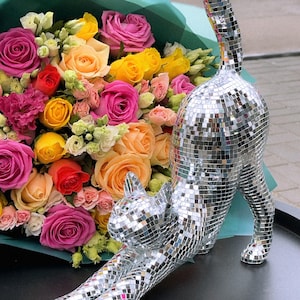 May include: A silver mosaic cat sculpture in a stretching pose, with a bouquet of colorful roses in the background. The roses are in shades of pink, yellow, orange, and red. The cat is made of small, reflective tiles.