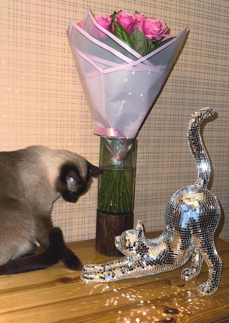 May include: A bouquet of pink roses in a clear vase, wrapped in white and pink paper, sits on a wooden surface. A Siamese cat and a decorative cat sculpture covered in mirrored tiles are also present.
