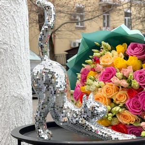 May include: A silver mosaic cat ornament in a stretching pose. The cat is covered in small, reflective squares. A bouquet of colourful roses in shades of pink, yellow, and orange is in the background.