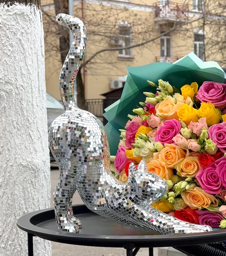 May include: A silver mosaic cat ornament in a stretching pose. The cat is covered in small, reflective squares. A bouquet of colourful roses in shades of pink, yellow, and orange is in the background.