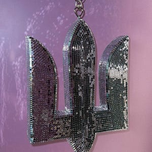 Puede incluir: Un tridente ucraniano de espejo plateado, un símbolo tradicional, cuelga de una cadena plateada. El tridente está hecho de pequeñas baldosas de espejo cuadradas.