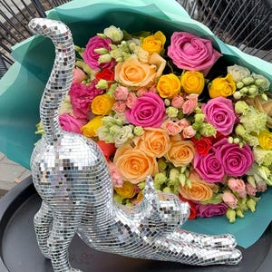 May include: A silver mosaic cat statue stretches out on a black surface, with a vibrant bouquet of roses in shades of pink, yellow, and orange in the background. The flowers are wrapped in a light blue paper.
