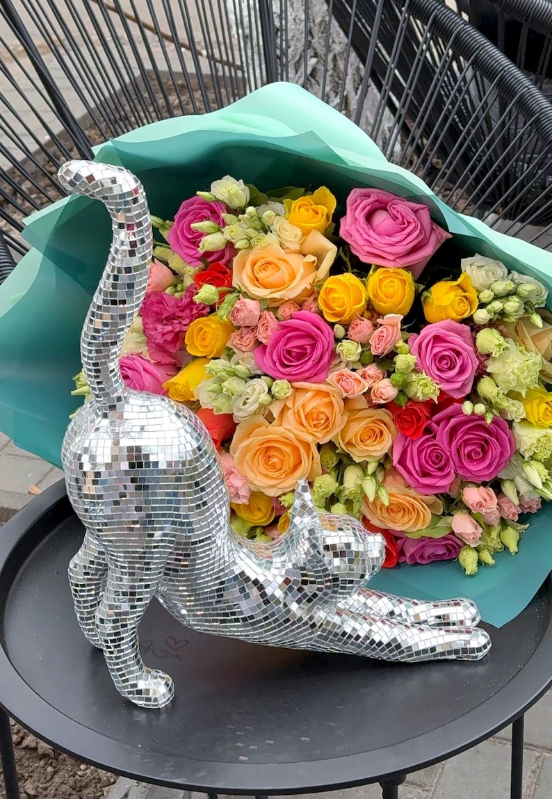 May include: A silver mosaic cat statue stretches out on a black surface, with a vibrant bouquet of roses in shades of pink, yellow, and orange in the background. The flowers are wrapped in a light blue paper.