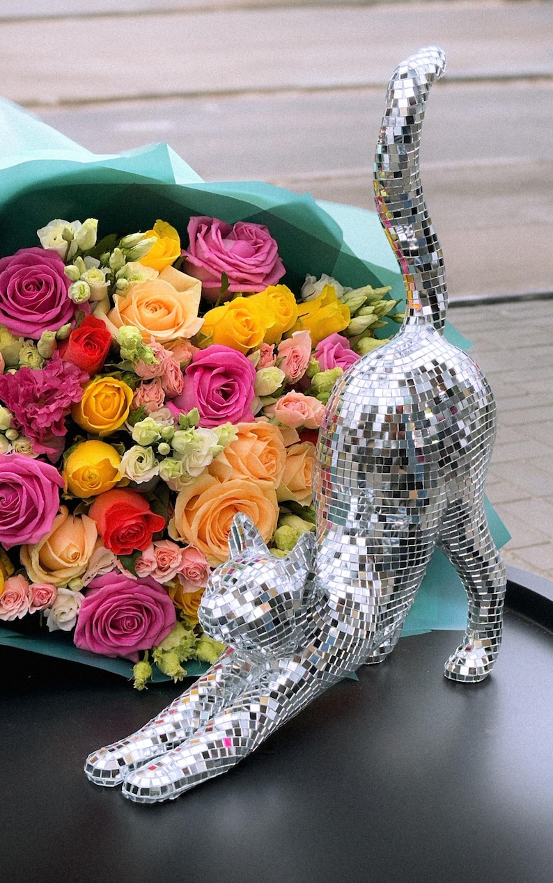 May include: A mosaic cat ornament in a stretching pose, with a bouquet of colourful roses in the background. The roses are in shades of pink, yellow, orange, and red. The cat is made of small, reflective tiles.