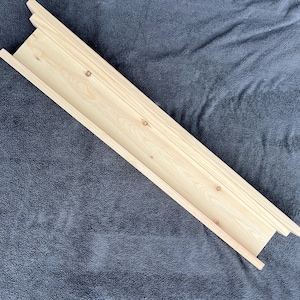 May include: A light-colored, unfinished wooden shelf with a rectangular design. The shelf has a shallow trough and is made of multiple wooden planks. The shelf is angled and rests on a dark gray textured surface.