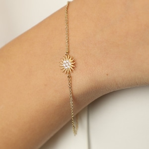May include: A delicate gold chain bracelet with a small, sparkling sun charm. The sun charm is made of gold and has a clear crystal center.