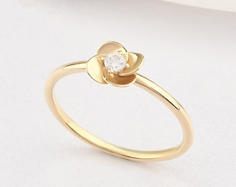 Può includere: Un delicato anello d'oro con un piccolo design a forma di fiore con una singola pietra trasparente al centro.