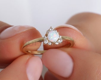 Puede incluir: Un anillo de oro con una piedra preciosa de ópalo en forma de pera, rodeada de pequeñas piedras claras. El anillo se sostiene entre dos manos, destacando el diseño. La banda del anillo es fina y elegante.