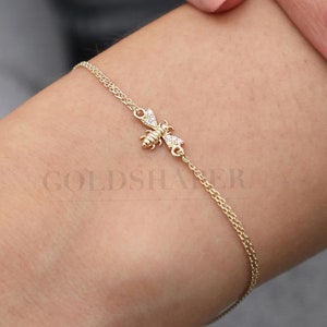 Könnte beinhalten: Ein zartes Armband aus einer goldenen Kette mit einem kleinen Bienenanhänger, der mit funkelnden, klaren Steinen verziert ist. Die Biene ist nach rechts gerichtet und hat ihre Flügel ausgebreitet.
