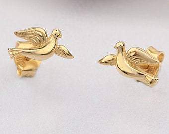 May include: A pair of gold-coloured dove stud earrings. Each earring features a detailed dove design with outstretched wings, showcasing a polished finish. The earrings are set against a white background, highlighting their metallic sheen.