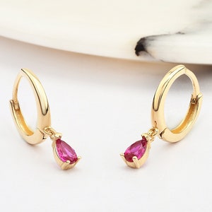 Könnte beinhalten: Ein Paar goldene Creolen mit einem baumelnden rosafarbenen tropfenförmigen Edelstein.