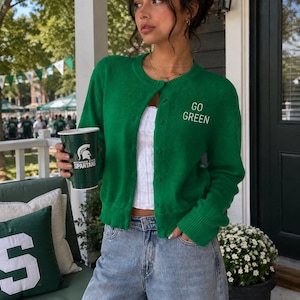 May include: A vibrant green long-sleeve button-down cardigan featuring the text GO GREEN embroidered in white on the upper left chest. The knit garment has a crew neckline, ribbed cuffs, and a ribbed hem, paired with light blue denim jeans.