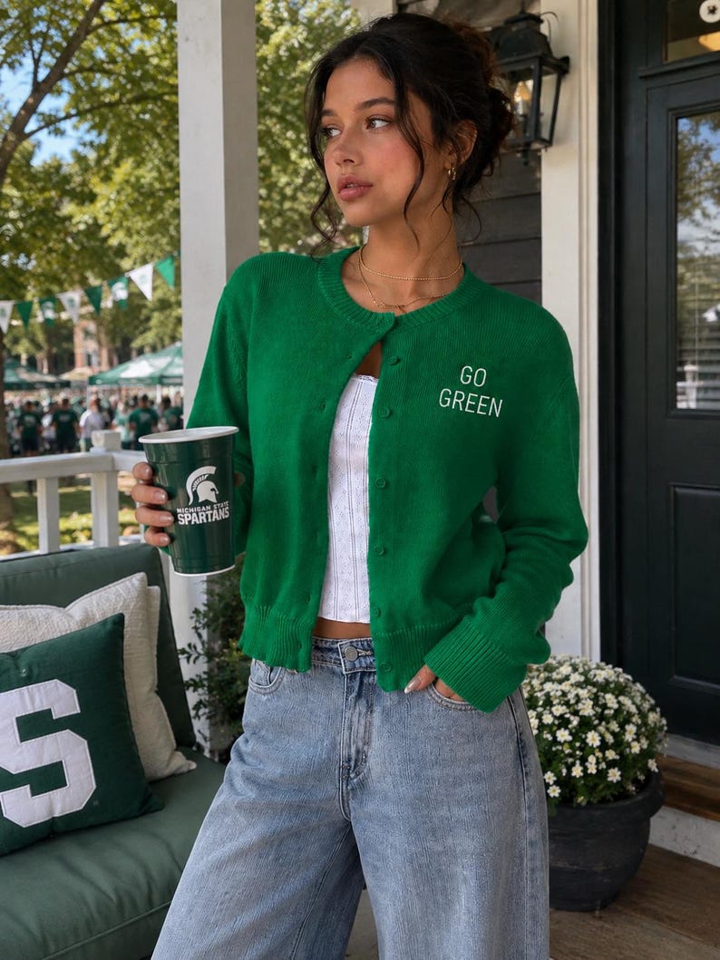 May include: A vibrant green long-sleeve button-down cardigan featuring the text GO GREEN embroidered in white on the upper left chest. The knit garment has a crew neckline, ribbed cuffs, and a ribbed hem, paired with light blue denim jeans.