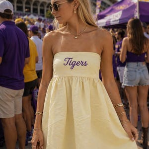 Op de afbeelding: Een lichtgeel strapless minijurkje met het woord Tigers in paars cursief schrift op de borst. Het kledingstuk heeft een aansluitend lijfje en een geplooide, uitlopende rok die boven de knie valt, gemaakt van een lichtgewicht stof.
