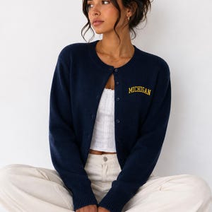 May include: A navy blue long-sleeve button-down cardigan featuring the word MICHIGAN embroidered in yellow thread on the upper left chest. The knit garment has a round neckline, ribbed cuffs, and a row of matching navy blue buttons down the front.
