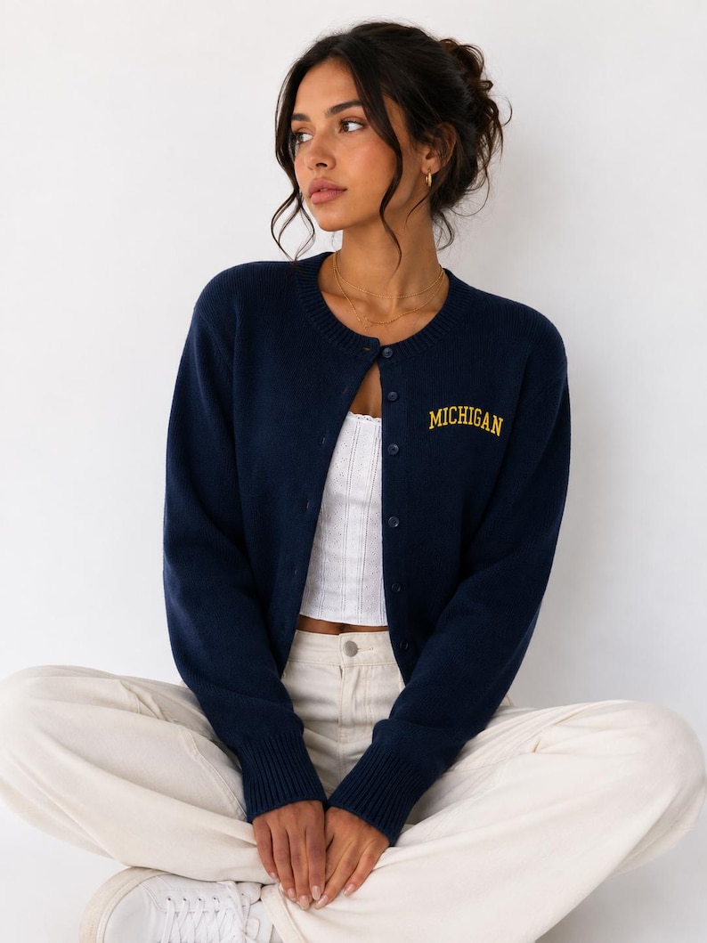 May include: A navy blue long-sleeve button-down cardigan featuring the word MICHIGAN embroidered in yellow thread on the upper left chest. The knit garment has a round neckline, ribbed cuffs, and a row of matching navy blue buttons down the front.