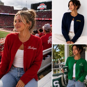 May include: A collection of three long-sleeve button-down cardigans in red, navy blue, and green. The red cardigan features the word Buckeyes, the navy blue one features the word Michigan, and the green one features the text Go Green.