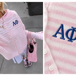 May include: A light pink and white striped hooded cardigan featuring a button-down front and the Greek letters Alpha Phi embroidered in dark blue on the left chest. The knit garment has long sleeves and a textured, soft appearance throughout.