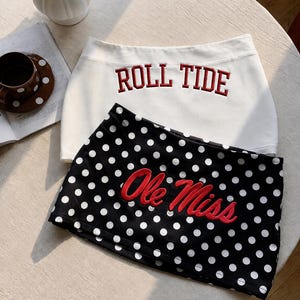 Pu&ograve; includere: Una gonna bianca con il testo ricamato in rosso ROLL TIDE e una gonna nera a pois bianchi con il testo ricamato in rosso Ole Miss. Entrambe le gonne presentano un orlo dritto e sono disposte sovrapposte su una superficie circolare.