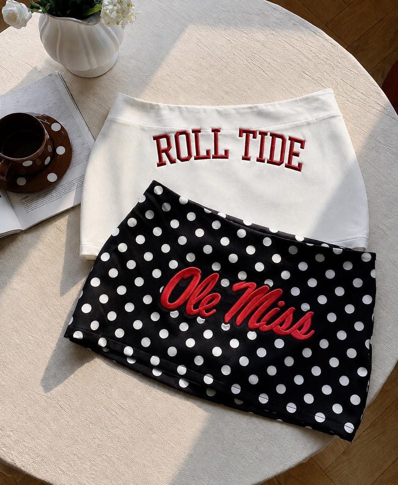 Pu&ograve; includere: Una gonna bianca con il testo ricamato in rosso ROLL TIDE e una gonna nera a pois bianchi con il testo ricamato in rosso Ole Miss. Entrambe le gonne presentano un orlo dritto e sono disposte sovrapposte su una superficie circolare.