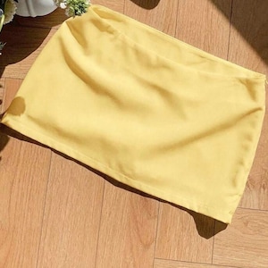 Pu&ograve; includere: Una minigonna giallo chiaro a tinta unita caratterizzata da una struttura in tessuto liscio e opaco, con un orlo dritto e pulito e una discreta chiusura a zip laterale, presentata in un design semplice con un colore pastello uniforme.