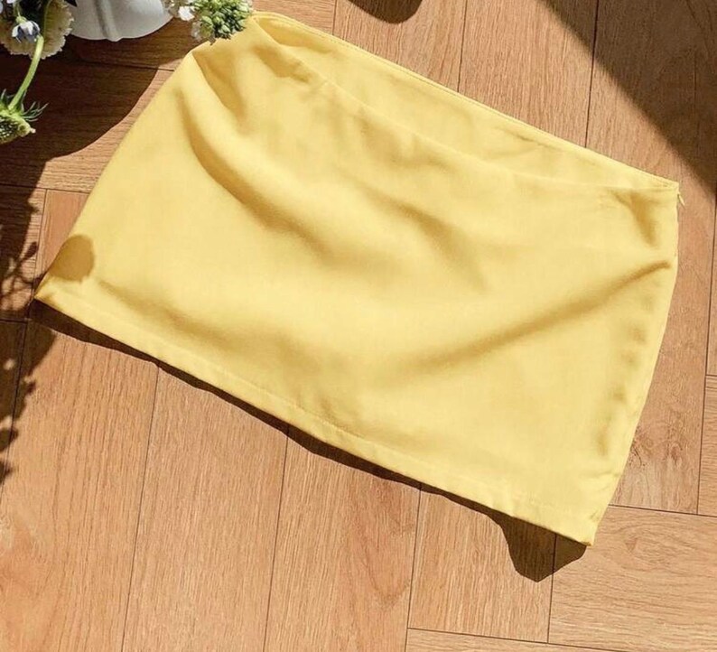 Pu&ograve; includere: Una minigonna giallo chiaro a tinta unita caratterizzata da una struttura in tessuto liscio e opaco, con un orlo dritto e pulito e una discreta chiusura a zip laterale, presentata in un design semplice con un colore pastello uniforme.